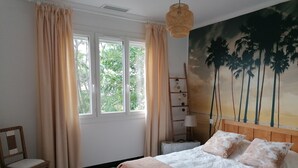 A bedroom with a bed, a chair, a window with curtains, a wall-mounted lamp, and a painting of palm trees. - GRANDE VILLA PROCHE MER WIFI PARKING (Agde)