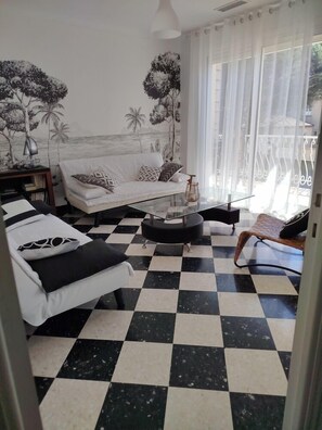 Uma sala de estar com piso xadrez, um sofá, uma mesa de centro e vista para o exterior através de cortinas finas.