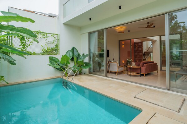 An indoor swimming pool with a glass wall, a seating area with a sofa and chairs, and a staircase leading to an upper level. - ELIVAAS Mirai Gemini 3 BHK Villa (Vagator)