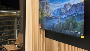 A modern hotel room with a large flat-screen TV mounted on a wooden wall, a sliding glass door leading to a balcony, and a foldable chair. - GH3-Glory Hieght- Vinhome Grand Park (Ho Chi Minh City)