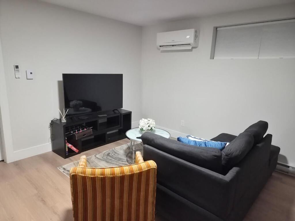 A living room with a black sofa, a striped armchair, a glass coffee table, a flat-screen TV, and an air conditioner mounted on the wall.
