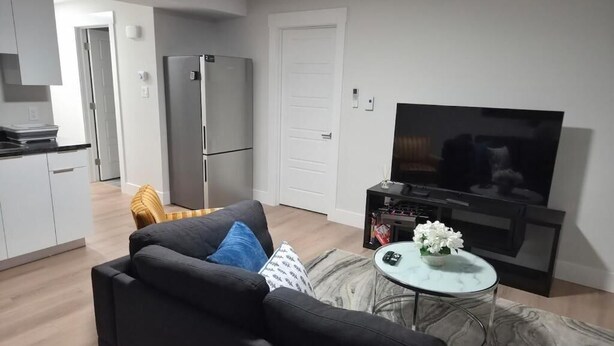Una sala de estar moderna con un sofá, un televisor de pantalla plana, una pequeña mesa de centro y una cocina al fondo.