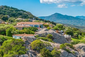 A villa with a tiled roof, surrounded by greenery and rocks, with a mountainous backdrop. - Elite Collection Pearl Villa - Luxury Private Spa (PORTO ROTONDO)