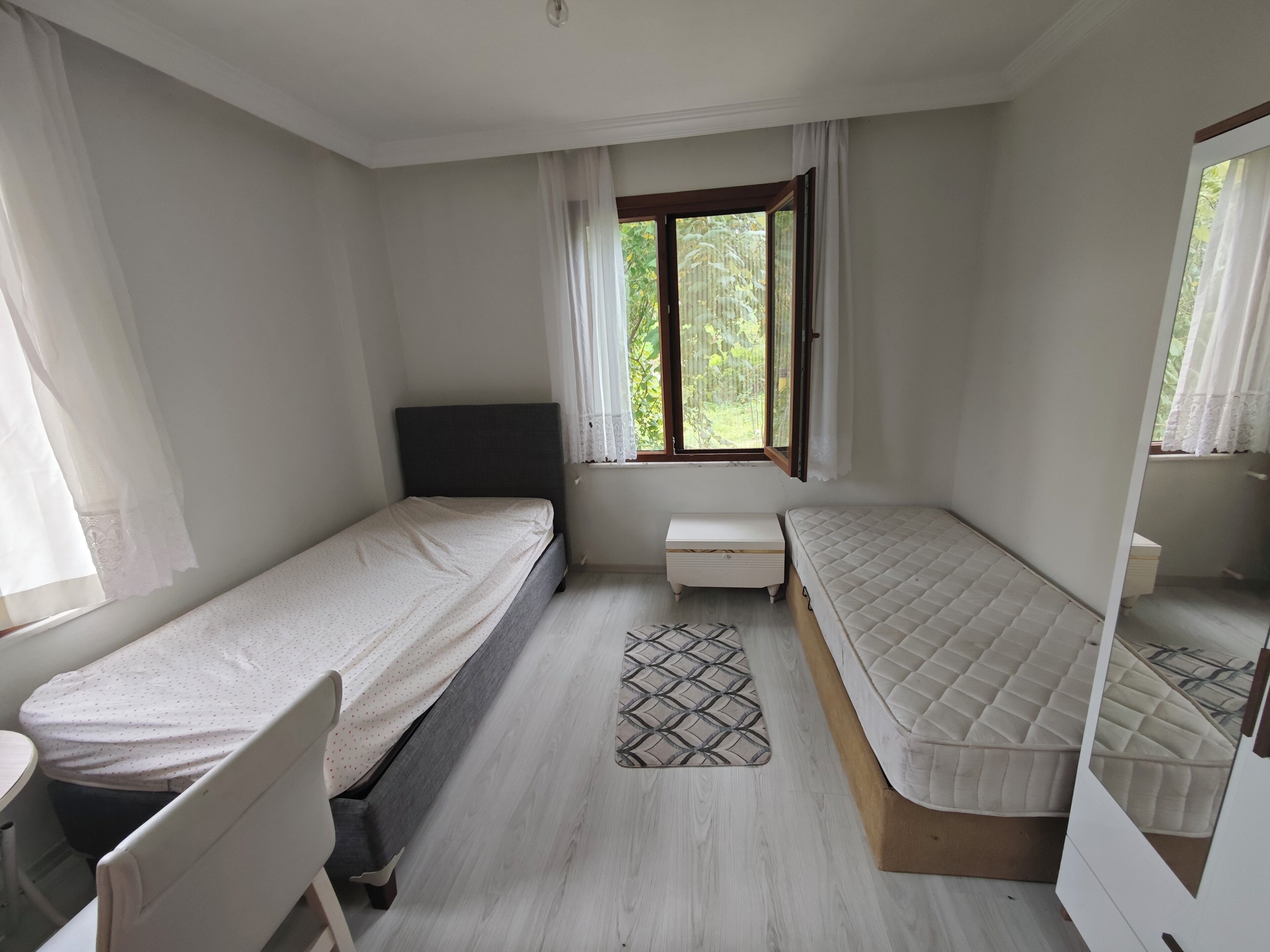 A bedroom with two single beds, a small bedside table, a chair, a window with curtains, and a patterned rug.