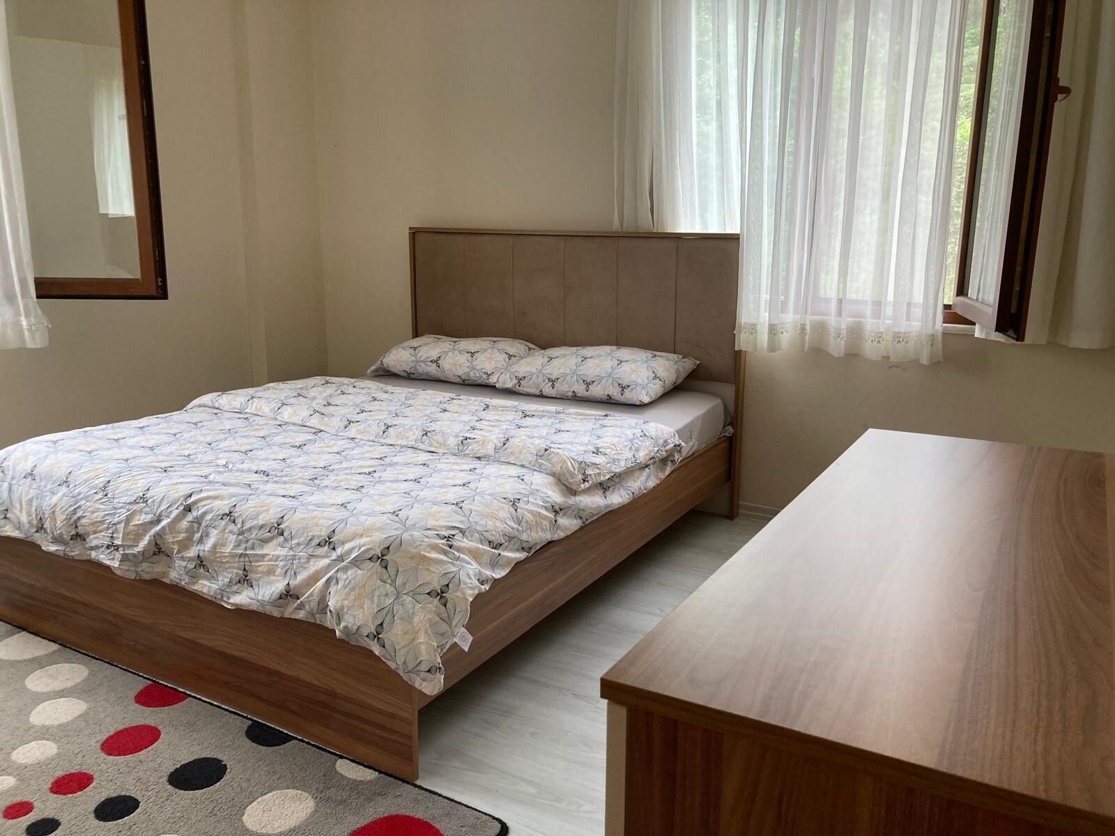 A bedroom with a bed, a wooden nightstand, a window with sheer curtains, and a patterned rug.
