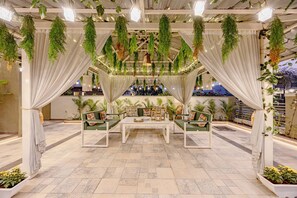 A covered outdoor seating area with a white table, green cushioned chairs, hanging plants, and potted plants on the floor. - ELIVAAS Heaven in Hills Mountara 3BHK  (Mavli)