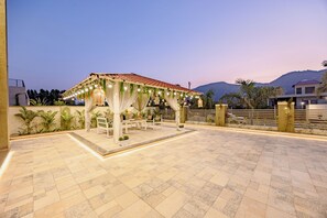 A covered outdoor seating area with a tiled floor, benches, and a pergola with string lights. - ELIVAAS Heaven in Hills Mountara 3BHK  (Mavli)