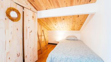 A small, wooden-paneled room with a sloped ceiling, a bed with a patterned blanket, a ladder, and a door with a decorative wreath.