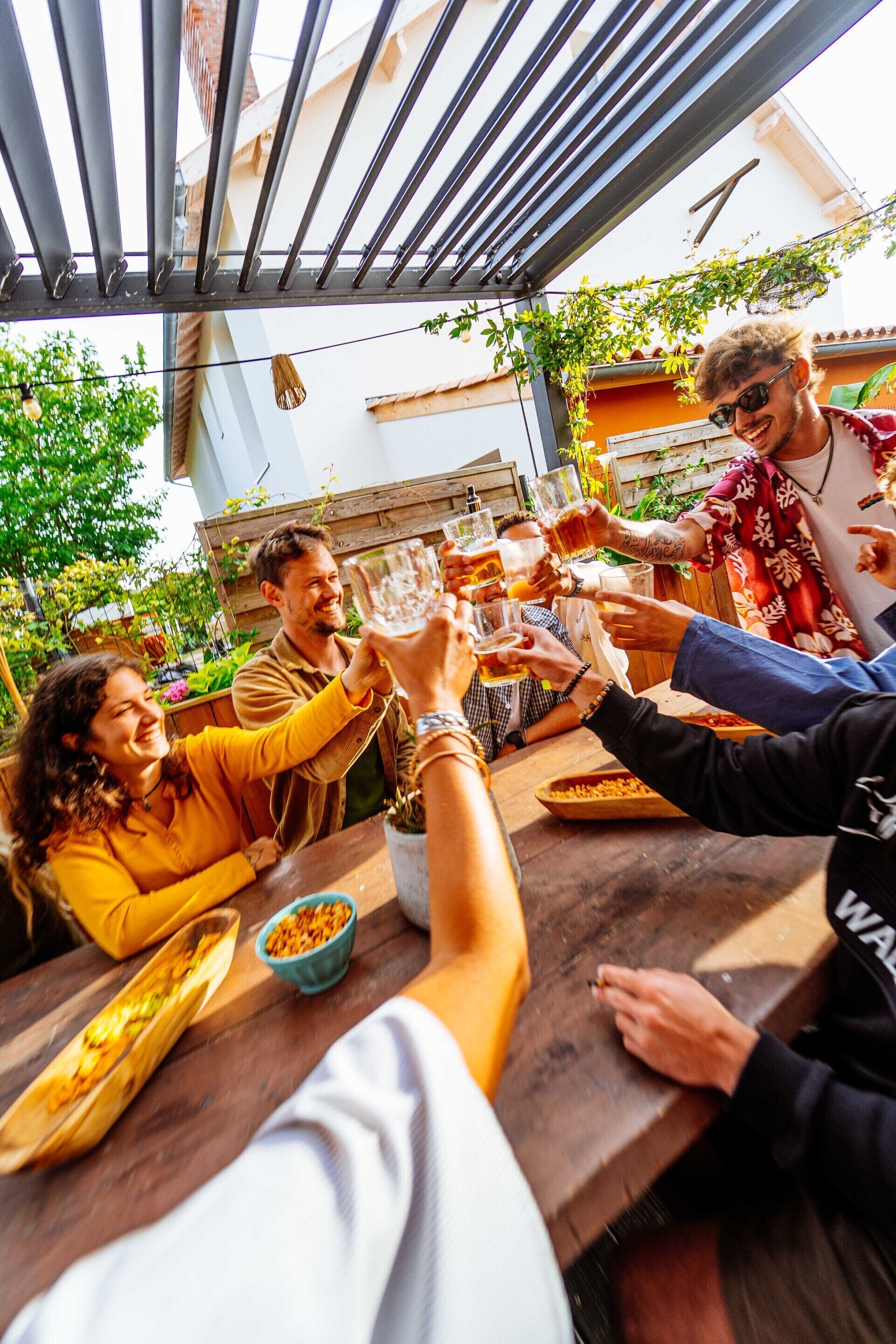A group of people toasting at a wooden table outdoors, with a metal railing and greenery in the background.