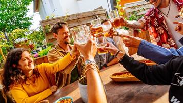 A group of people toasting at a wooden table outdoors, with a metal railing and greenery in the background.