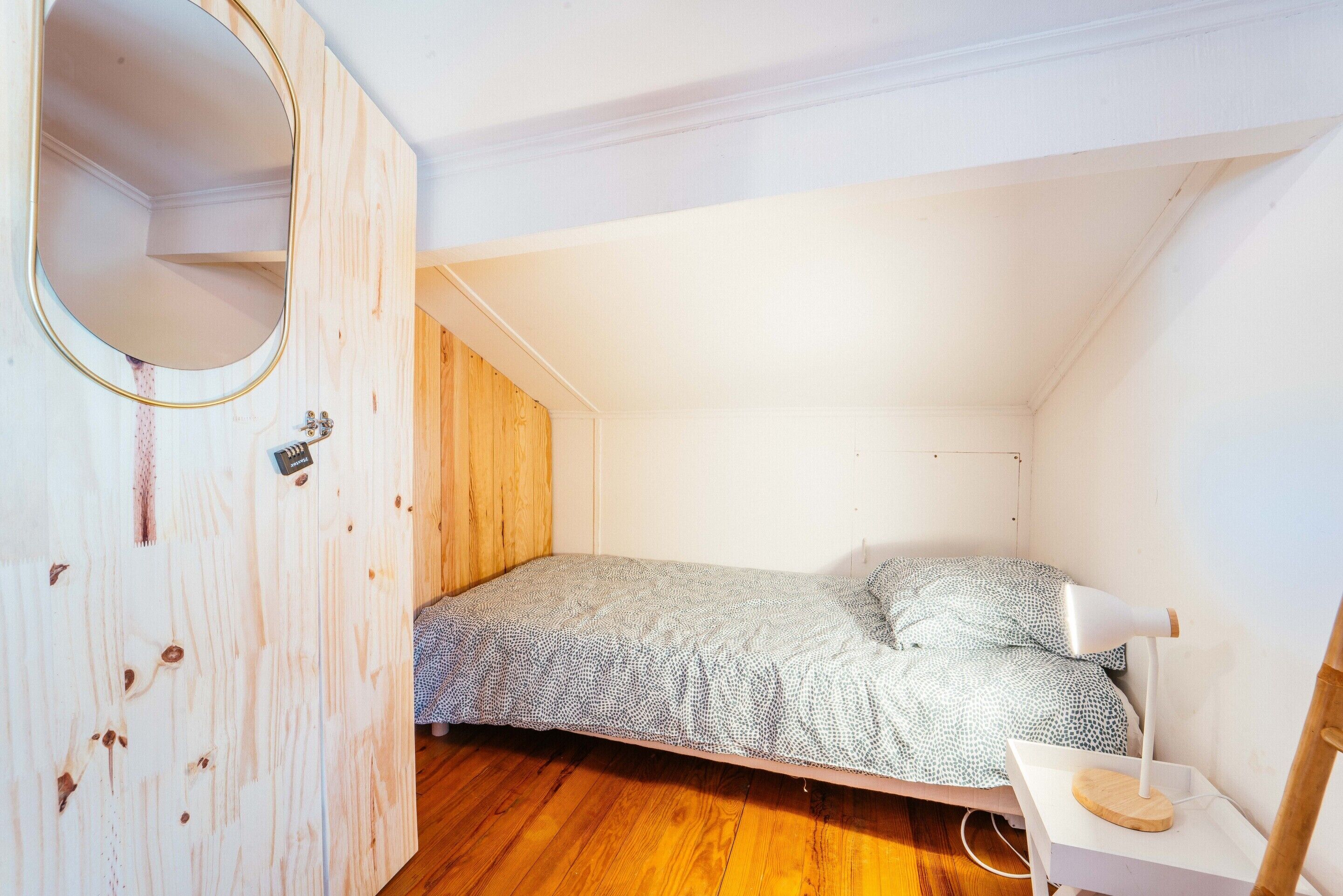 Une petite chambre avec un lit simple, un plafond en pente, une armoire en bois, une table de chevet avec une lampe et une couvre-lit à motifs.
