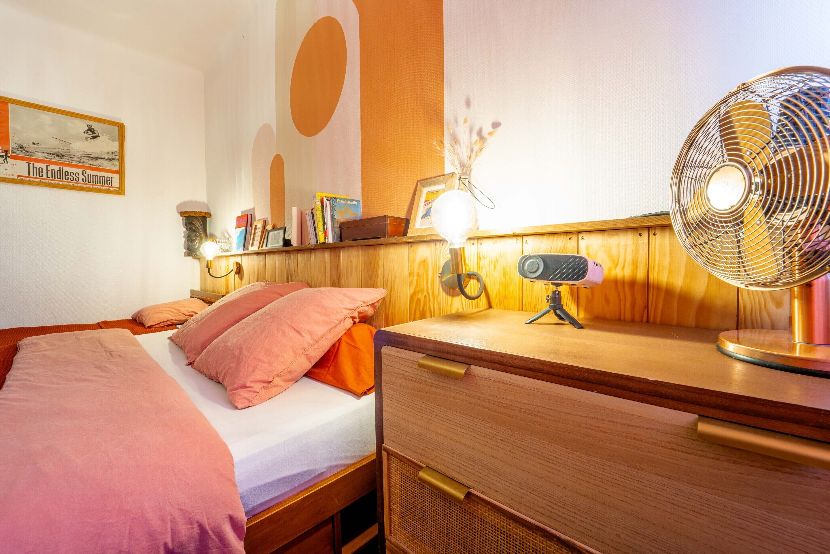 Une chambre avec un lit en bois, une table de chevet avec une lampe, un bureau avec un ventilateur et un mur orné d’une affiche.
