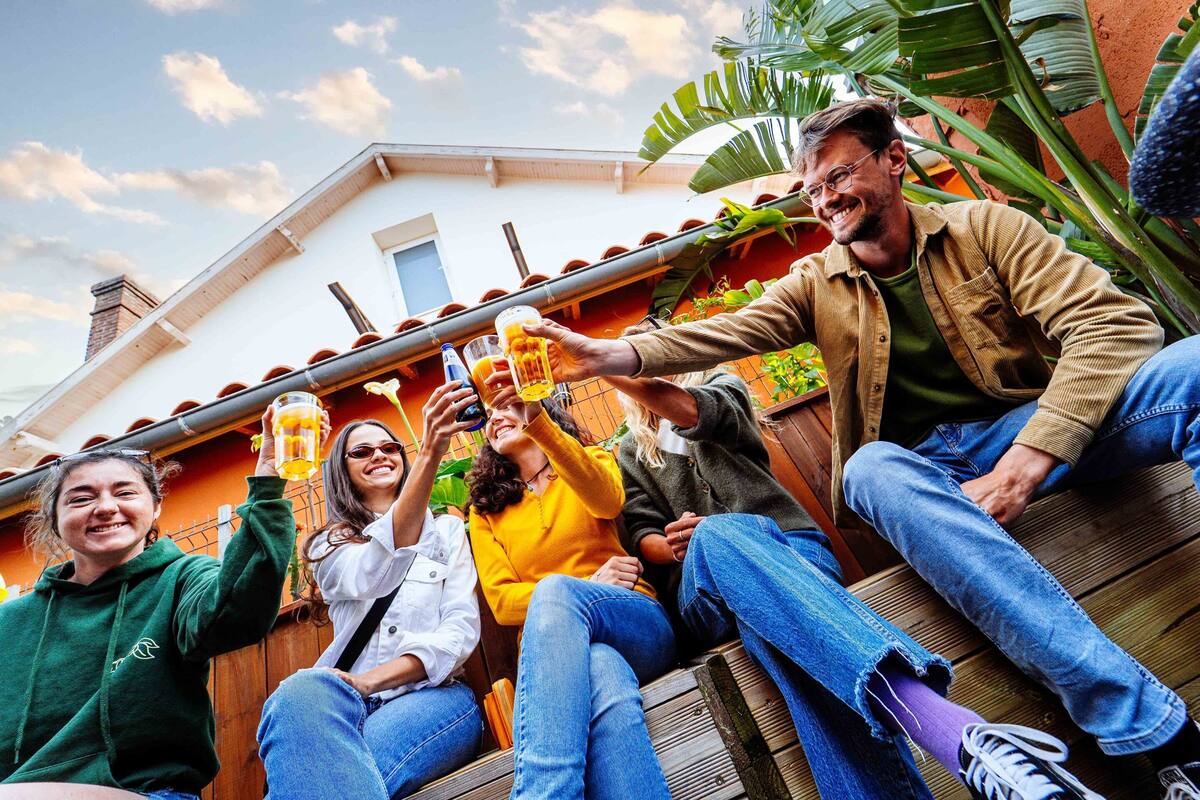 A group of people sitting on a wooden deck, holding beer mugs, with a house and plants in the background.