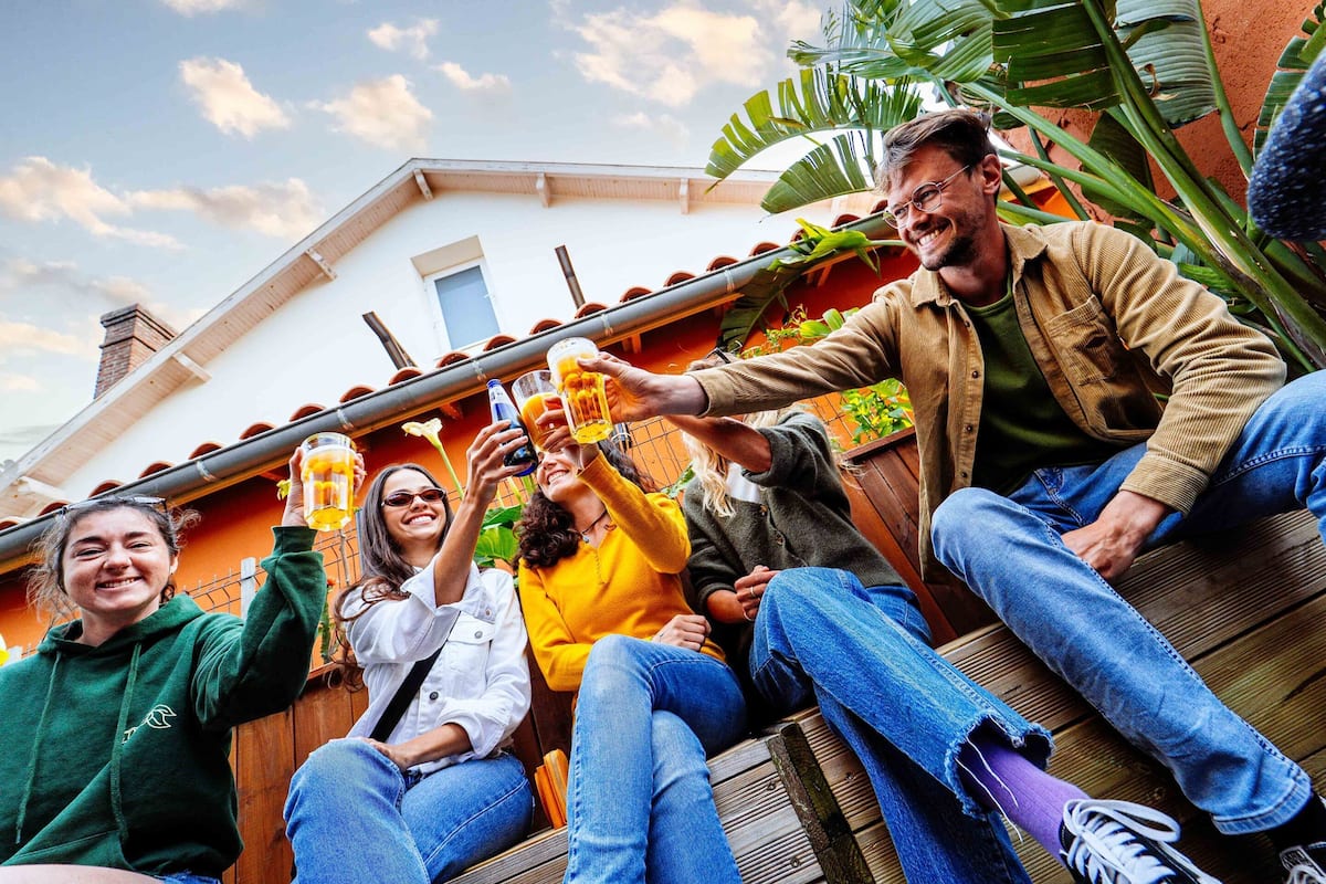 Un grupo de personas sentadas en una terraza de madera, sosteniendo jarras de cerveza, con una casa y plantas al fondo.
