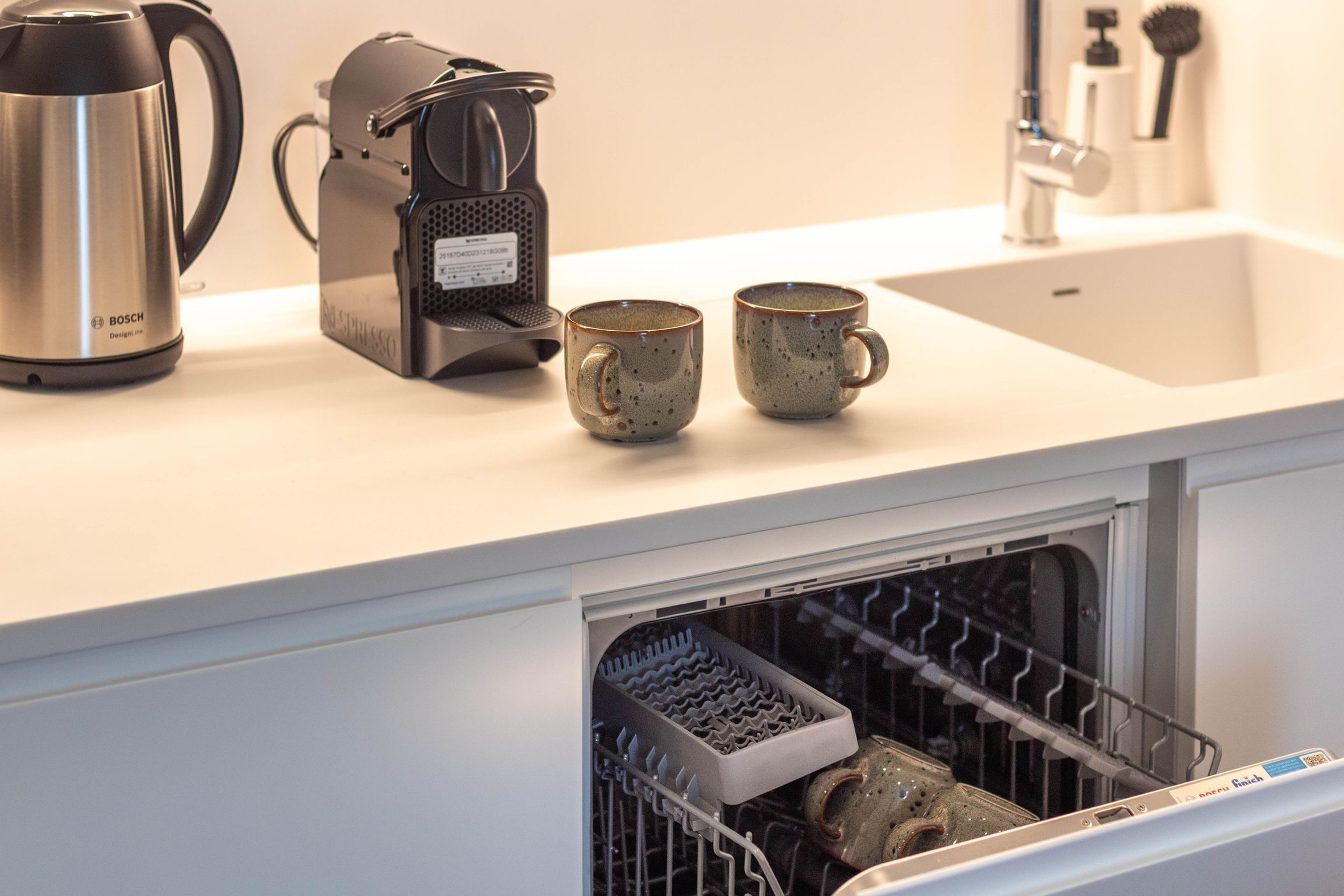 Una encimera de cocina con una cafetera Bosch, dos tazas de cerámica y un lavavajillas con platos en su interior.