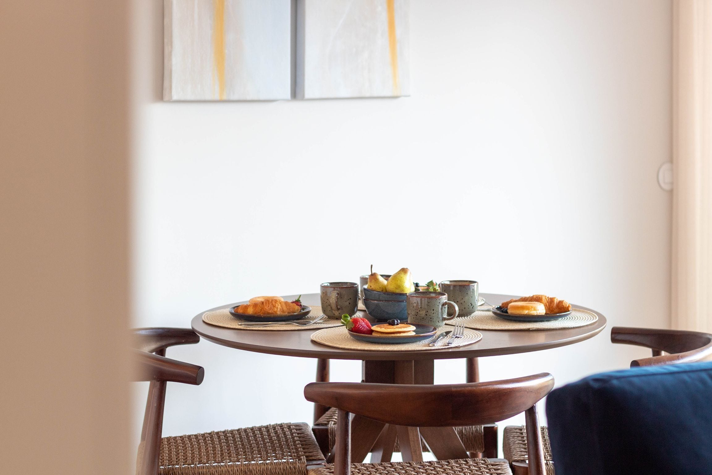 A round dining table with a plate of croissants, a bowl of fruit, and a cup. There are two chairs with woven seats and dark wood frames.