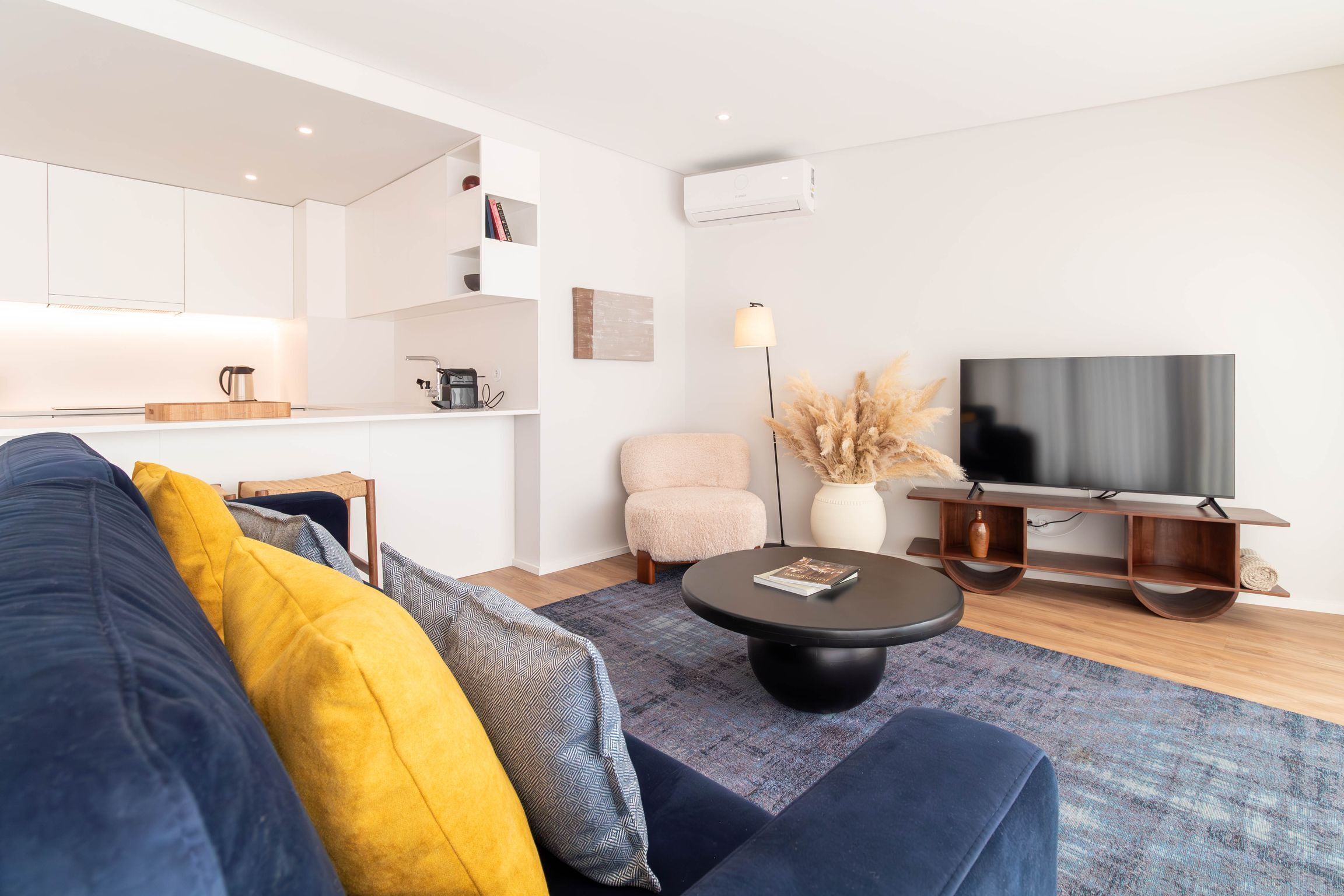 Una sala de estar moderna con sofá, mesa de centro, televisor y una zona de cocina con cafetera.
