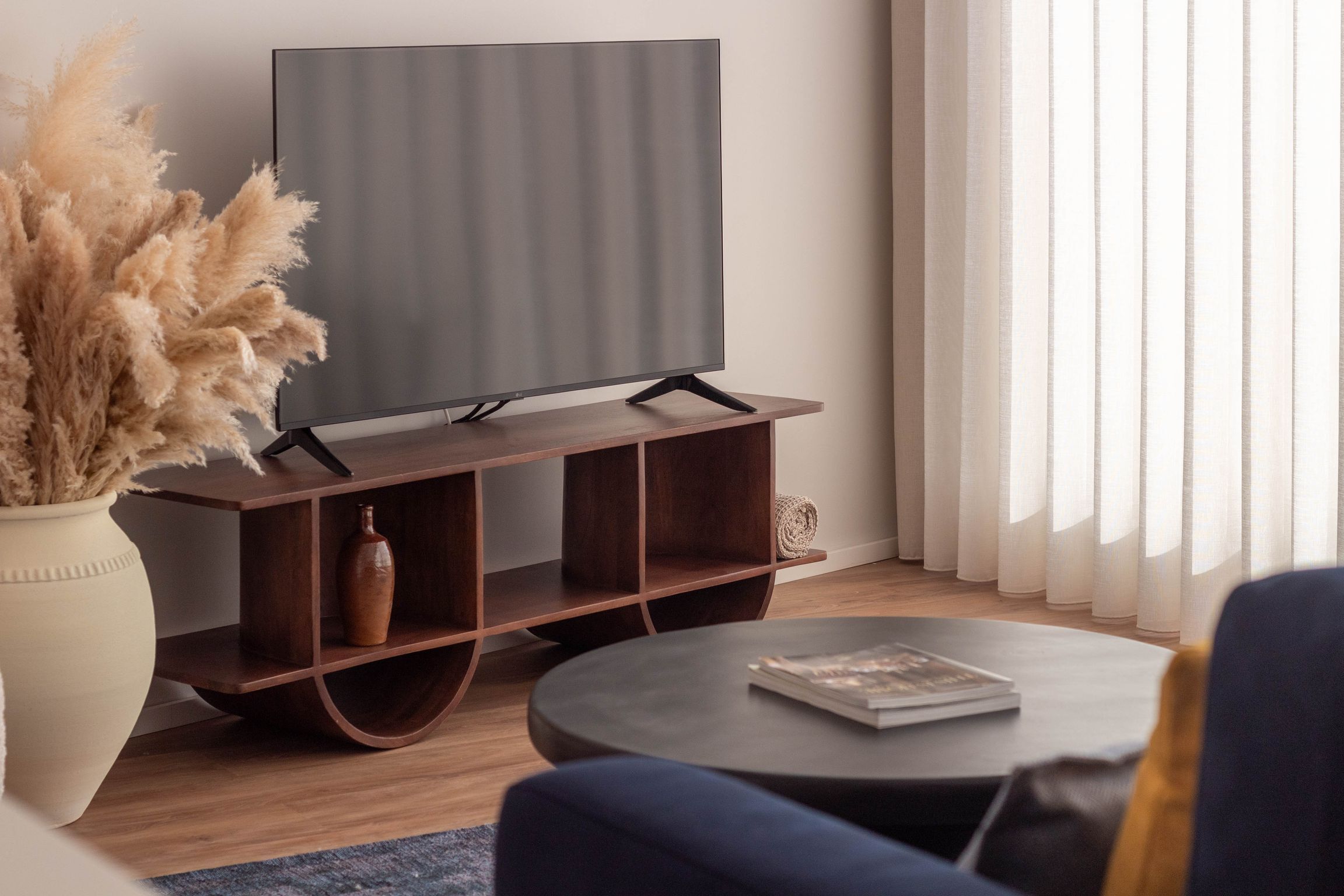 Una sala de estar moderna con un televisor de pantalla plana sobre un soporte de madera, una mesa de centro redonda con una revista y un jarrón con plantas secas.