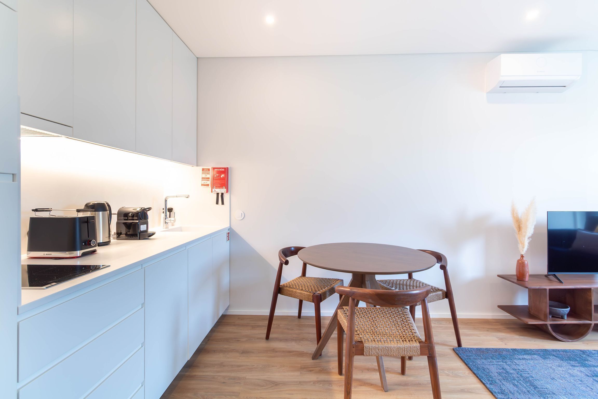 Uma cozinha moderna com uma cafeteira Bosch, micro-ondas e torradeira. Há uma área de refeições com uma mesa redonda e duas cadeiras de vime.