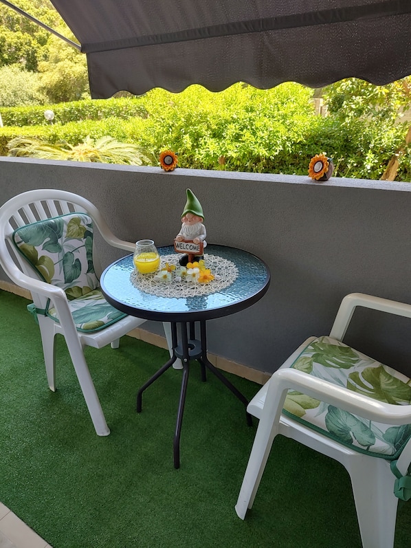 Une petite table ronde avec un plateau en verre, deux chaises blanches avec des coussins verts, un gnome de jardin tenant une pancarte, et un décor de verdure en arrière-plan.
