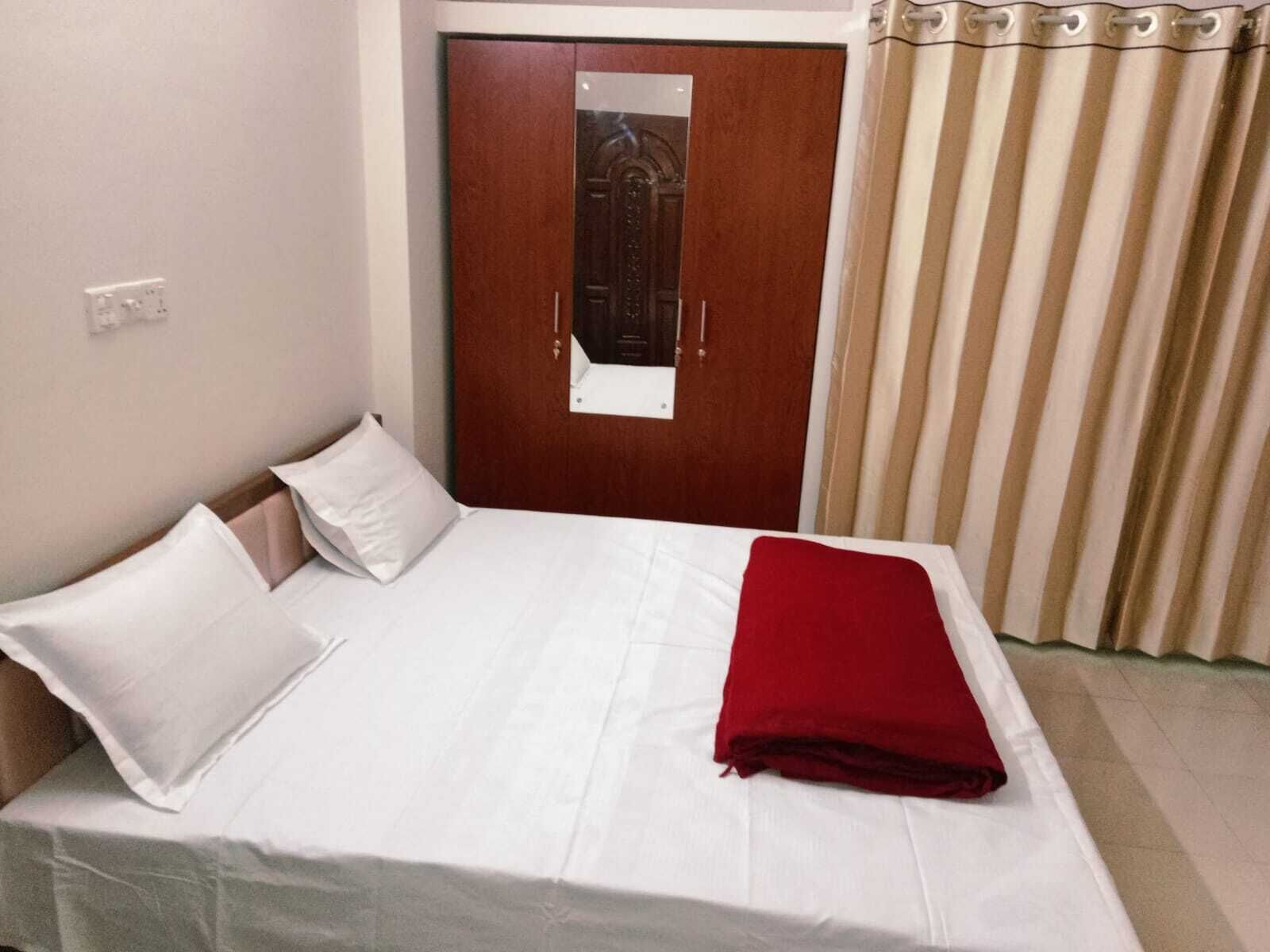 A neatly made bed with white bedding and a red blanket, a wooden door with a mirror, and beige curtains.
