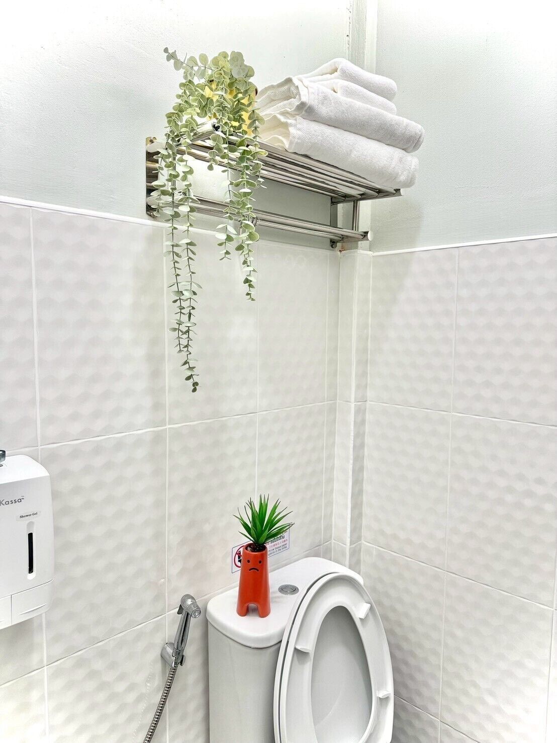 Un baño con inodoro, secamanos, repisa con toallas y una planta colgante.