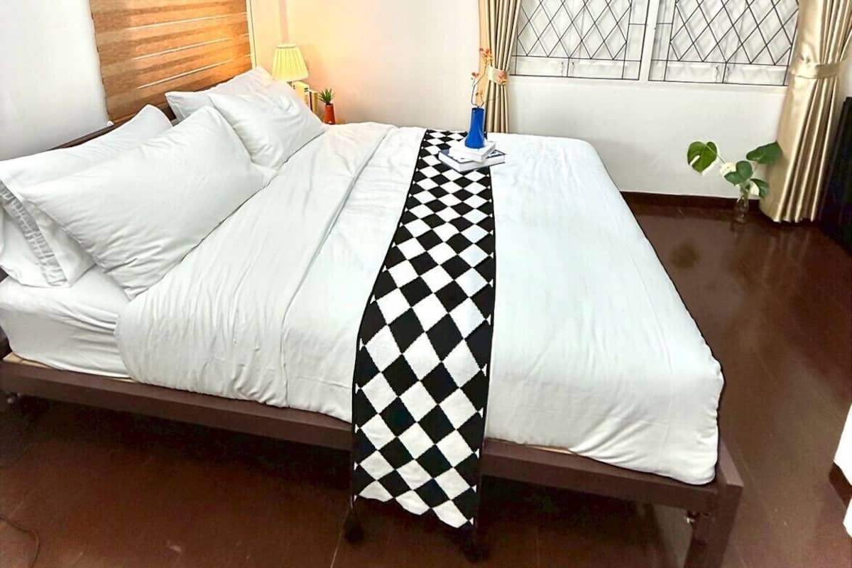 A hotel room with a bed, a nightstand, a window with curtains, a framed picture on the wall, and a potted plant.