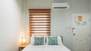 Habitación de hotel con cama, mesita de noche con lámpara, ventana con persianas, unidad de aire acondicionado y puerta con tablón de anuncios.