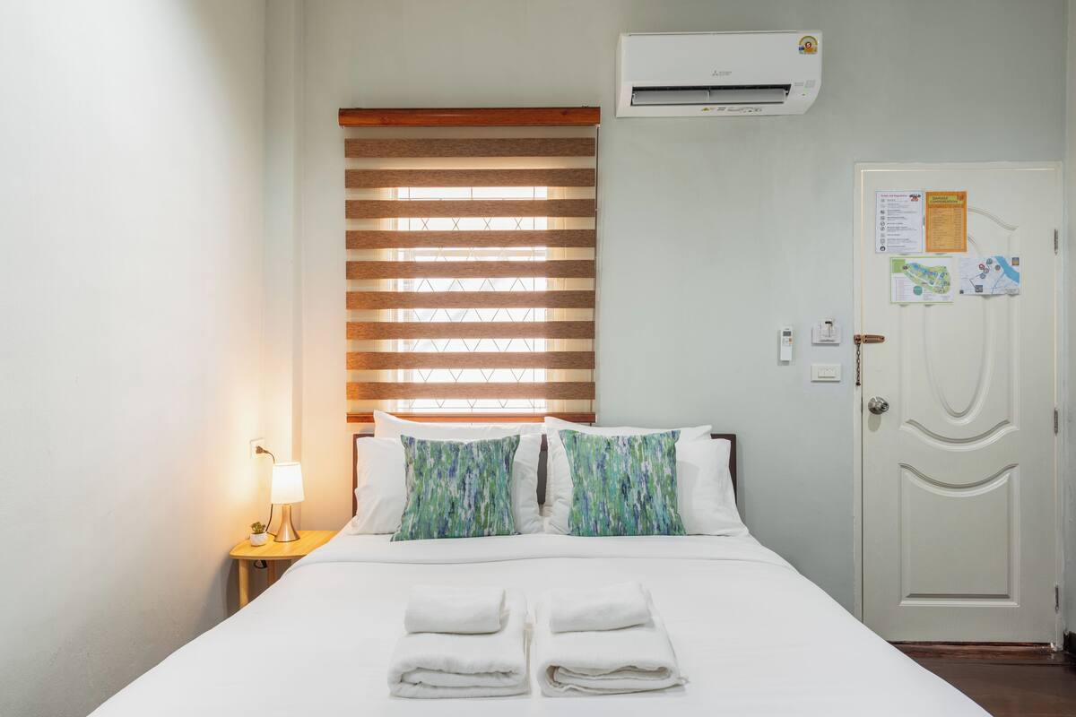 A hotel room with a bed, a nightstand with a lamp, a window with blinds, an air conditioning unit, and a door with a bulletin board.