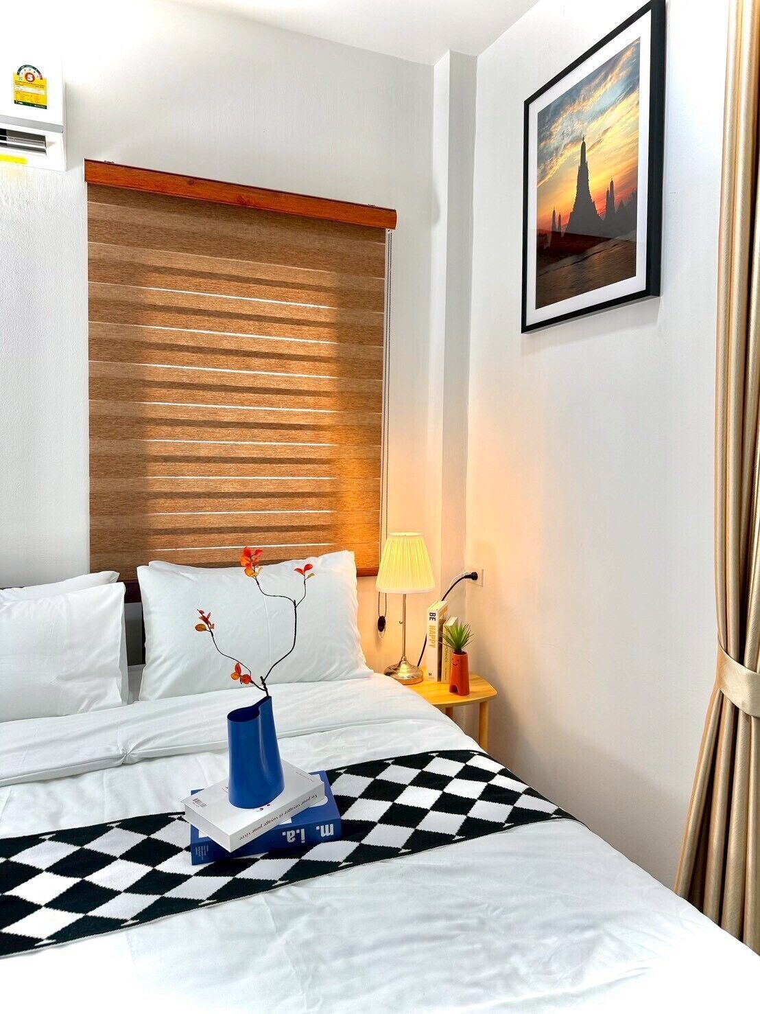 A hotel room with a bed, a nightstand, a lamp, a framed picture on the wall, and a window with blinds.