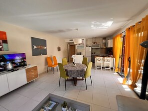 A modern living room with a flat-screen TV, a dining area with patterned tablecloth, and a kitchen with orange curtains. - Apartment Cavalaire center near beaches 75 m2 garden floor beautiful terrace (Cavalaire-sur-Mer)