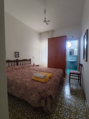 A bedroom with a bed, a wooden wardrobe, a chair, a ceiling fan, and a bathroom visible through an open door.