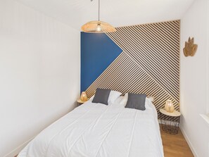 A bedroom with a bed, a hanging light fixture, a wall with a geometric pattern, and a small table with a lamp. - 600m to the beach – House for 6 with veranda (Treffiagat)