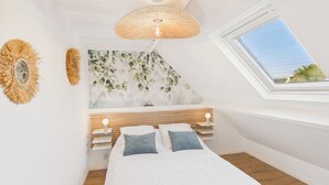 Un dormitorio con techo inclinado, tragaluz, cama con ropa de cama blanca y pared con estampado de hojas.