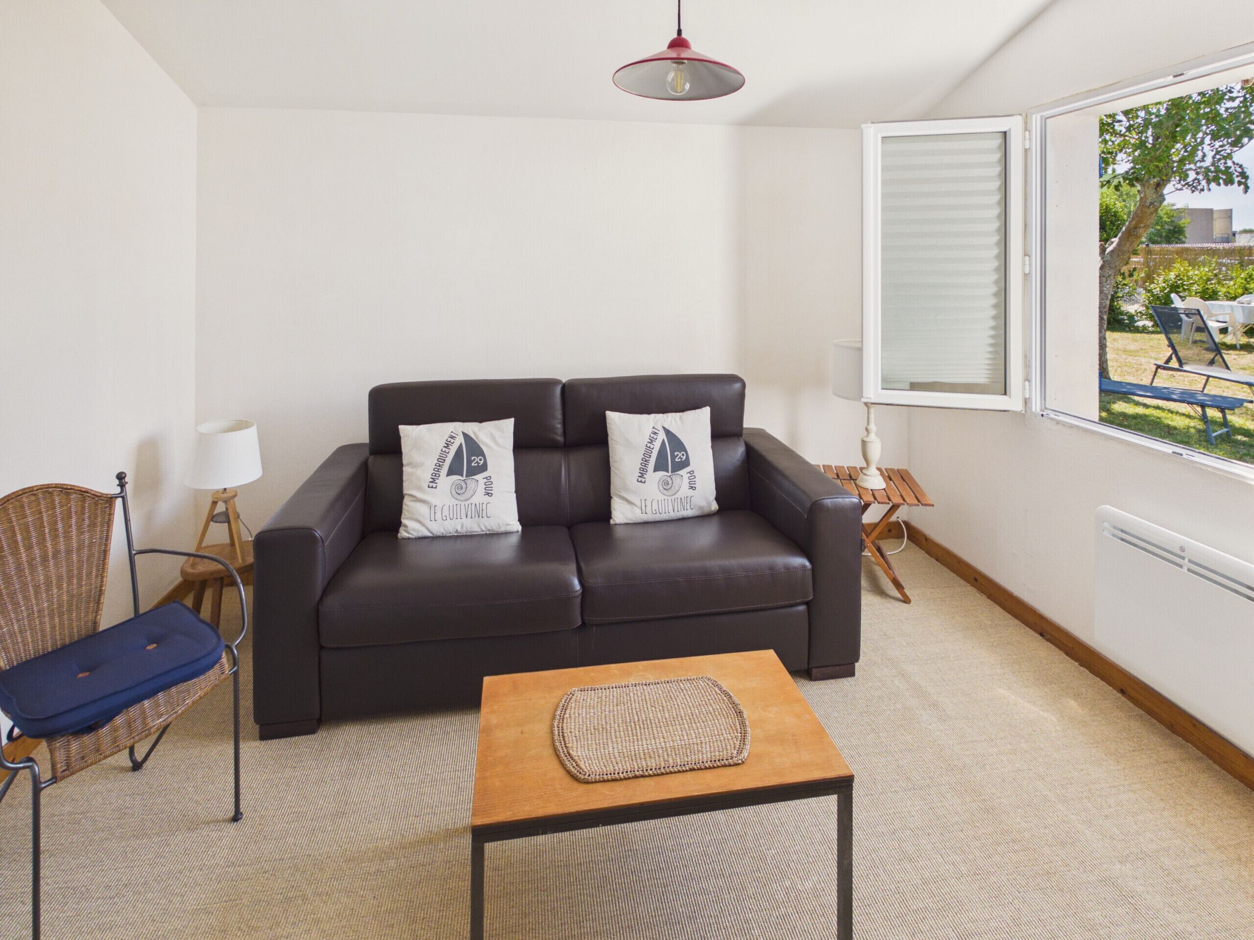 A living room with a dark leather sofa, two matching pillows, a wicker chair, a small wooden table, and a view through a window.