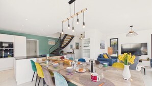 A modern kitchen with white cabinets, a dining area with colorful chairs, a wooden dining table, and a staircase leading to an upper level. - Superb recently built house for 8 with garden (Pléneuf-Val-André)