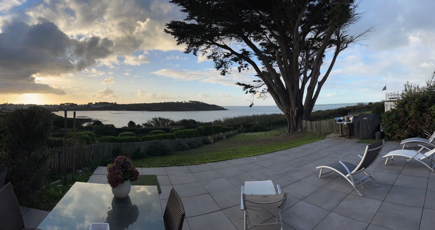 Une terrasse extérieure avec une table et des chaises, un grand arbre et une vue sur la mer.