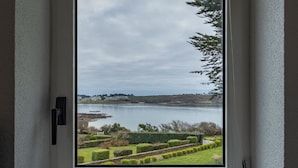 A window with a view of a lake, greenery, and distant buildings. - House Ocean Front Arzon Port Navalo (Arzon)