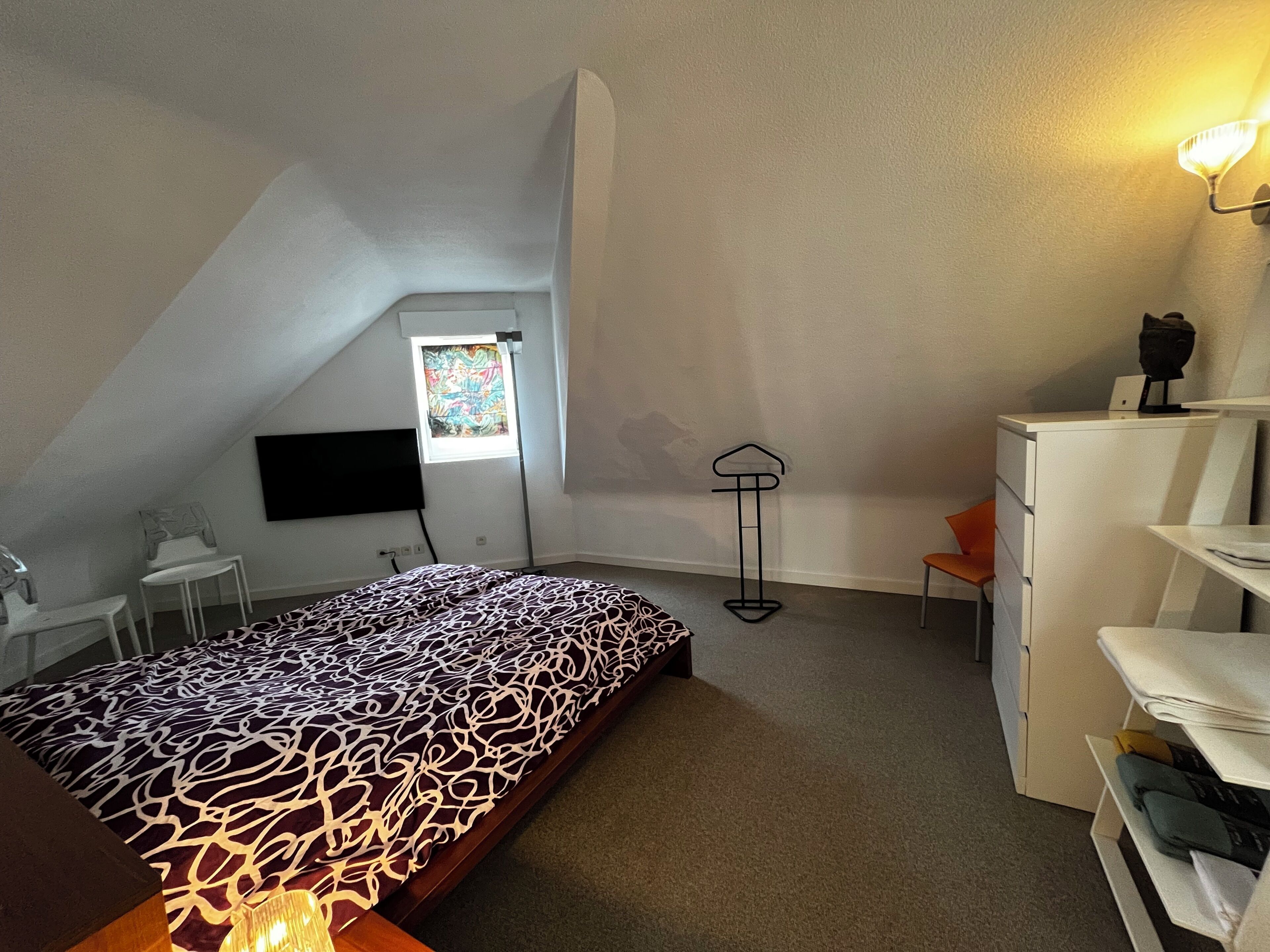 A bedroom with a bed, a desk, a chair, a TV, and a window.