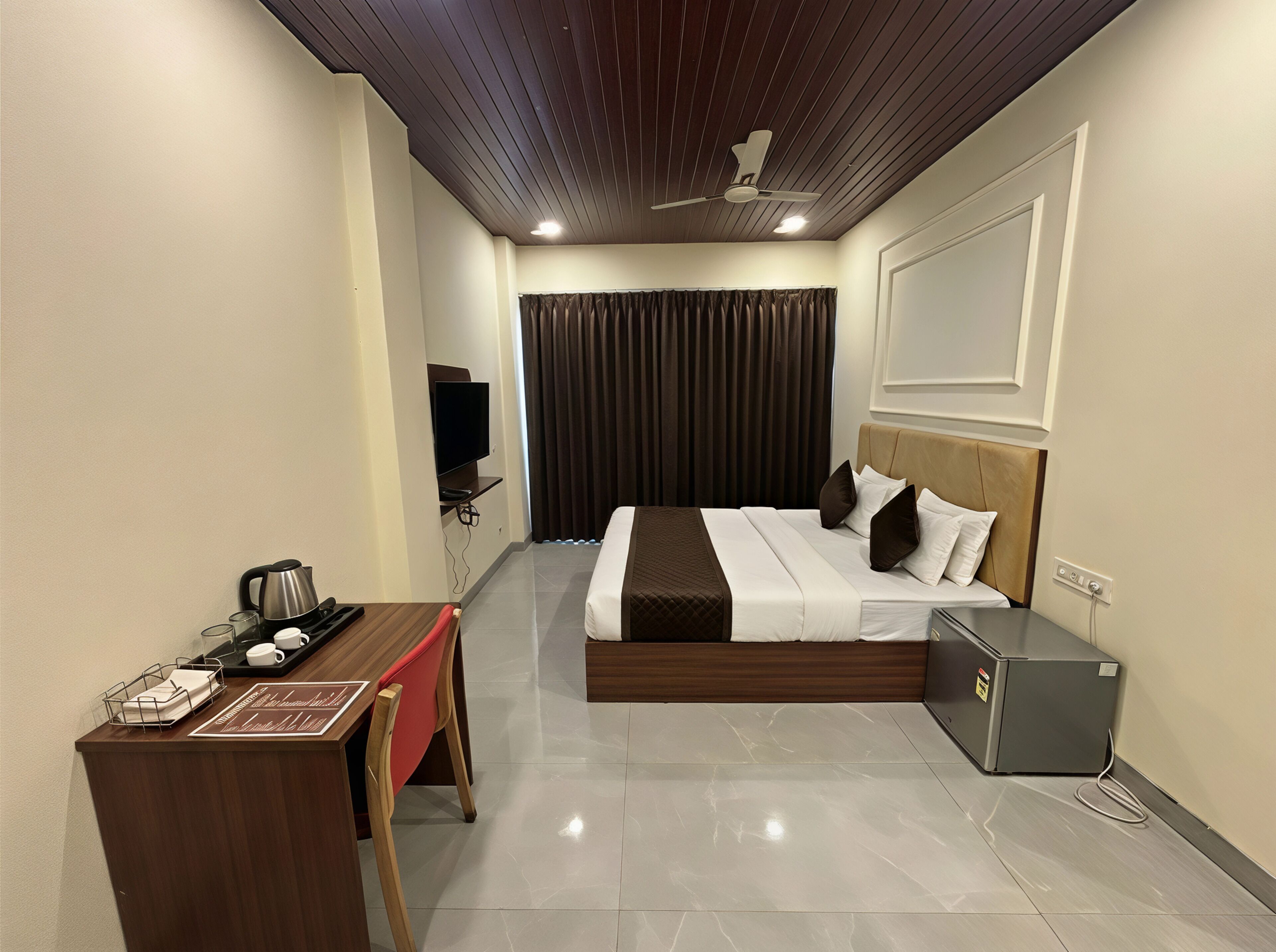A hotel room with a bed, a desk with a kettle, a television, and a refrigerator.
