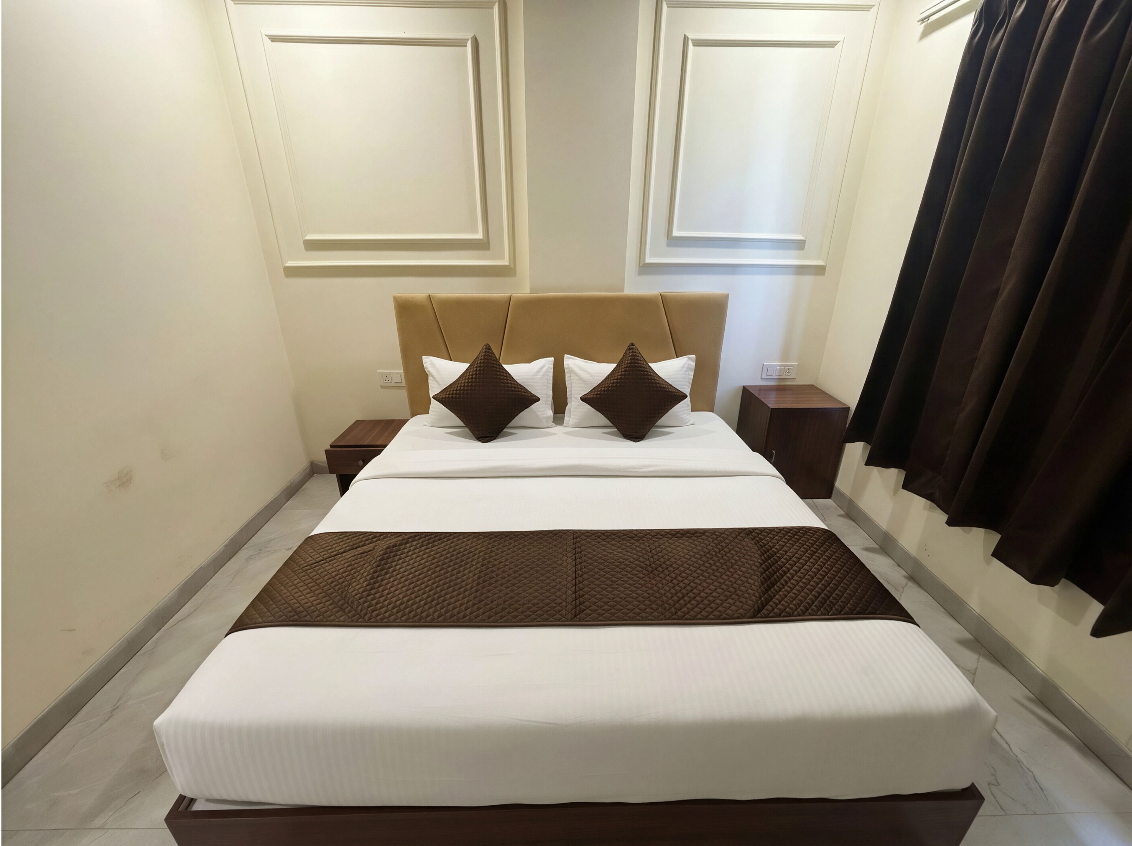 A neatly made bed with a brown quilt, white pillows, and a headboard. There is a nightstand with a lamp and a power outlet. The room has a tiled floor and a dark curtain on the window.