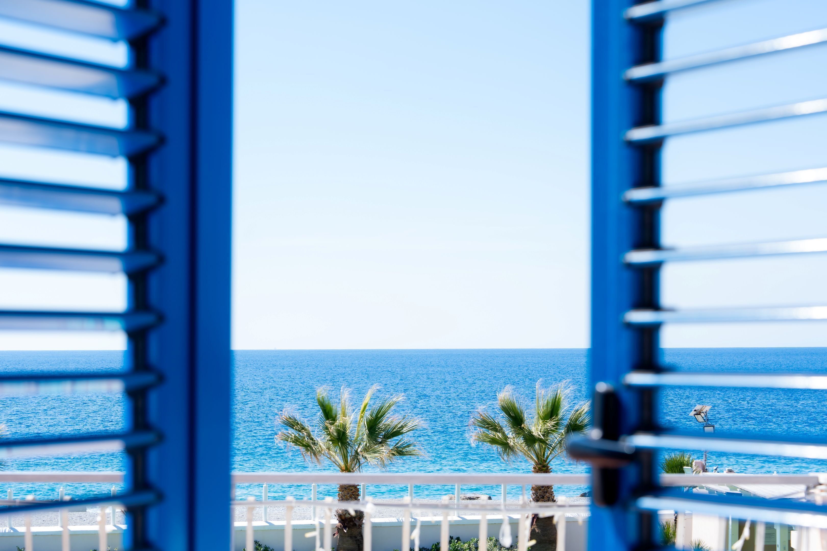 A view through a window with blue shutters, overlooking a beach with palm trees and a clear blue sea.