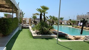 A pool area with artificial turf, palm trees, lounge chairs, and a clear blue sky. - Didimos Resort (Montebello Ionico)