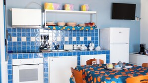 A kitchen with blue tiles, a white refrigerator, a table with orange tablecloth, and a TV on the wall. - Didimos Resort (Montebello Ionico)