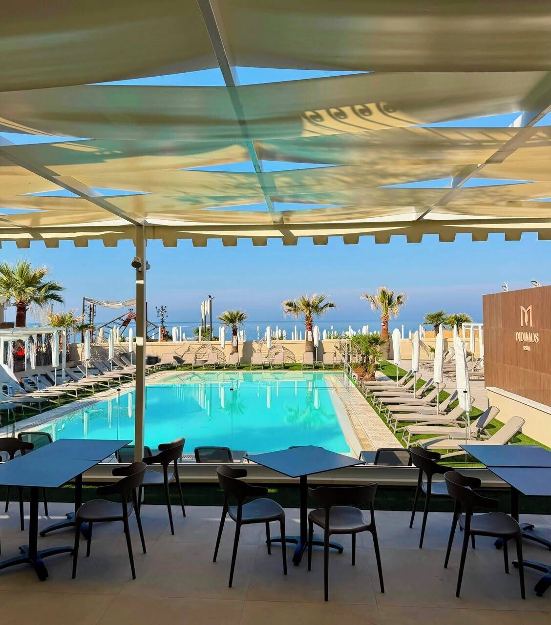 A pool area with lounge chairs and tables, a clear view of the sea, and a building with a sign.