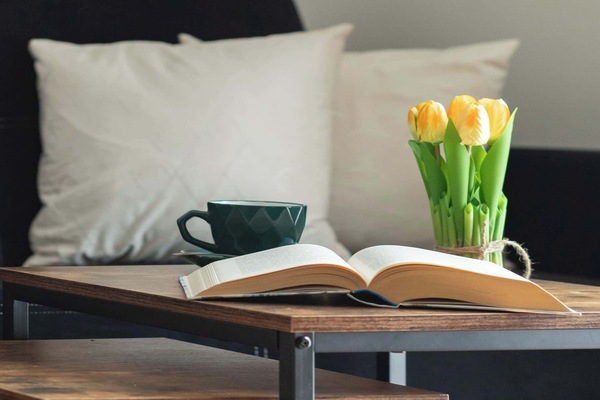 A close-up shot focuses on a wooden side table decorated with a white pillow, an open book, and a vase of yellow tulips. This small nook creates a perfect spot for a morning coffee.