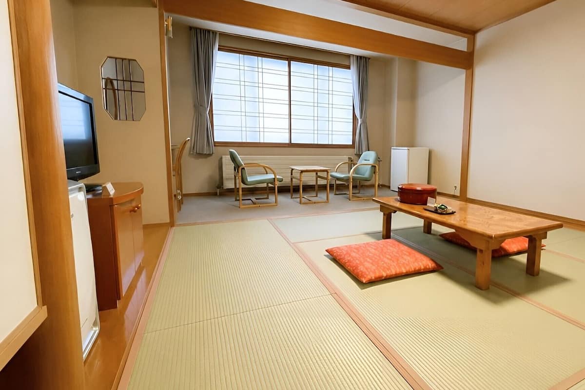 A traditional Japanese room with tatami flooring, a low table, chairs, a television, and a window with curtains.