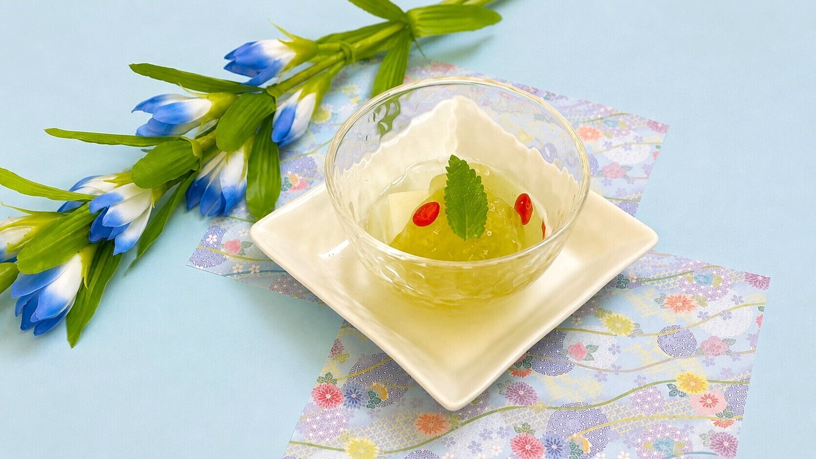 A clear glass bowl with a yellow liquid, a mint leaf, and red chili slices on a floral-patterned napkin.