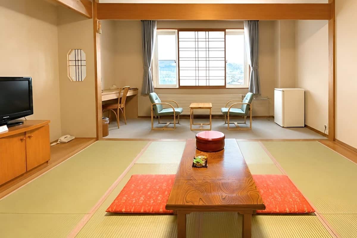 A traditional Japanese tatami room with a tatami mat floor, a wooden table with a red cushion, two chairs, a television on a cabinet, and a window with curtains.