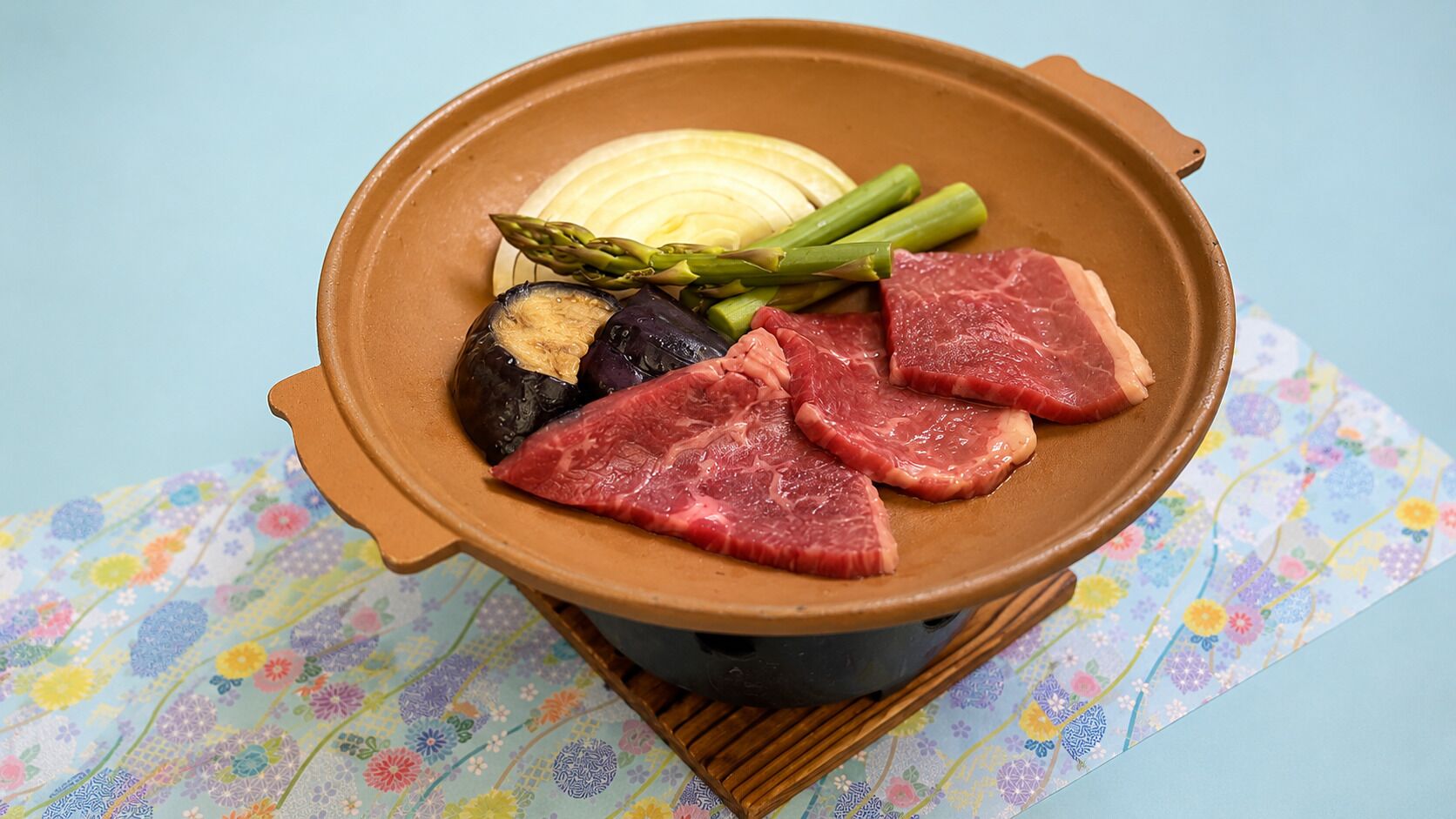 A hot pot with meat, asparagus, and eggplant on a colorful tablecloth.